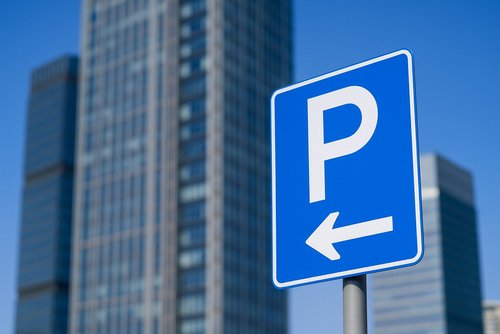 Blaues Parkplatzschild mit Pfeil nach links vor modernen Hochhäusern bei klarem Himmel.